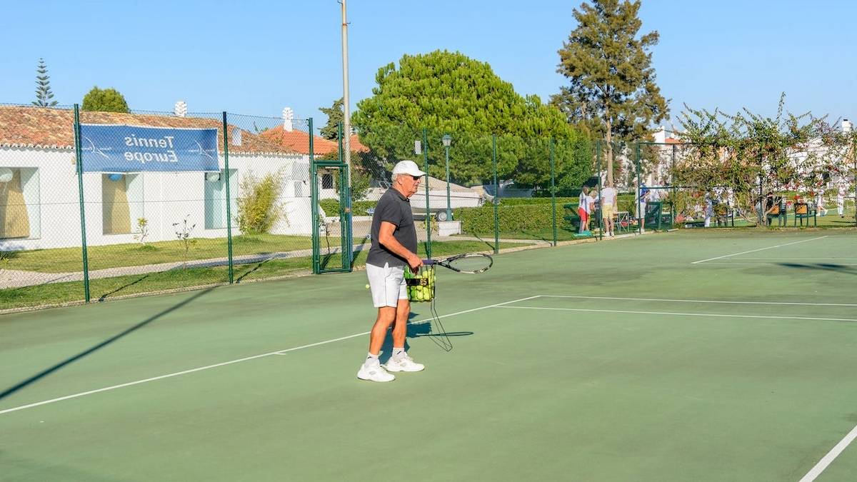 Tennisplatz im Pedras da Rainha Resort mit einem Mann, der Tennisschläger und Ballkorb hält.