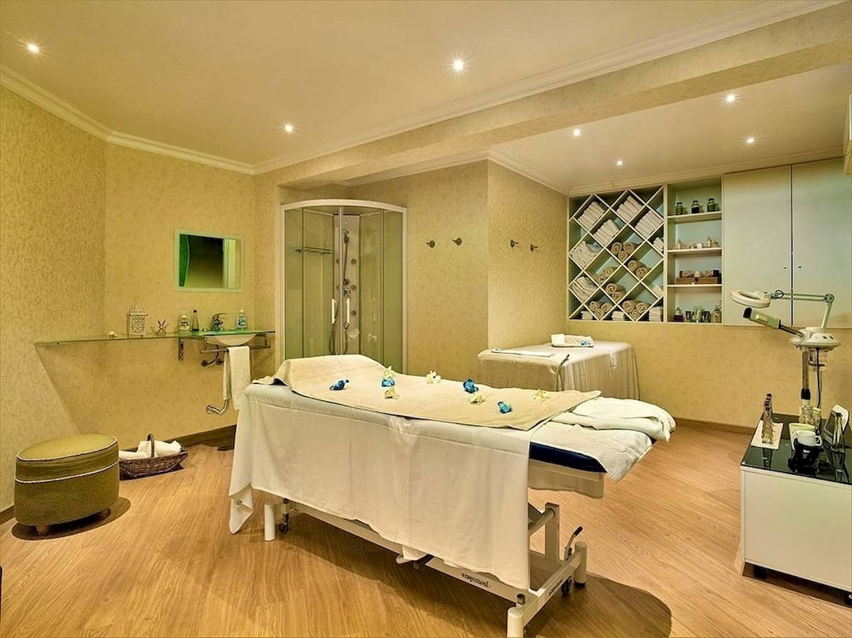 Behandlungsraum im Baia Grande Hotel mit zwei Behandlungstischen, einer Dusche und Regalen mit Wellnessartikeln.