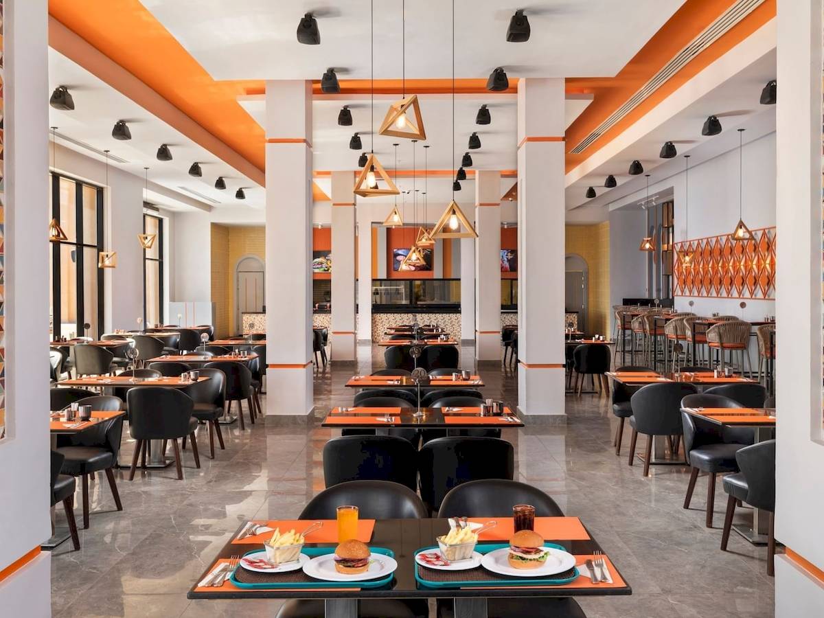 Innenansicht des Restaurants im Pickalbatros Water Valley Resort by Neverland mit runden Tischen, schwarzen Stühlen und orangen Tischdecken.