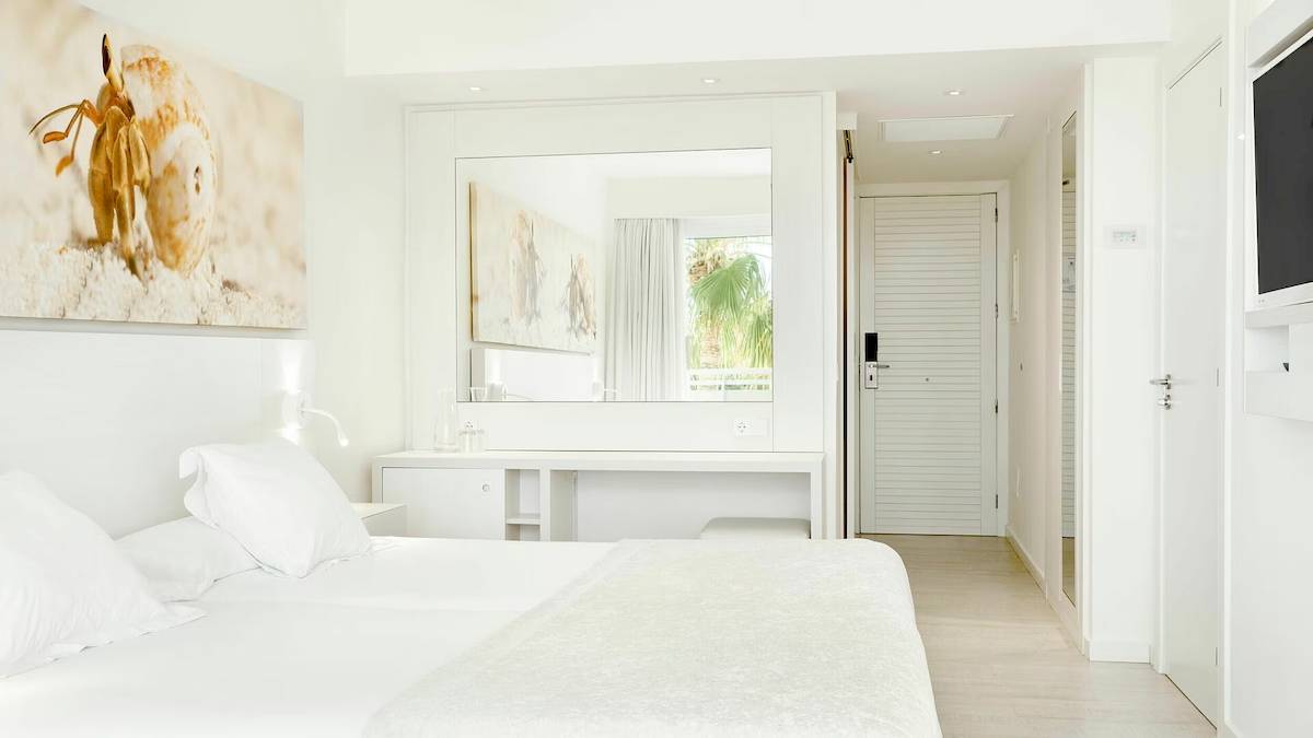 Innenansicht eines Schlafzimmers im Iberostar Cristina Hotel mit weißem Bett, Möbeln und Dekoration.