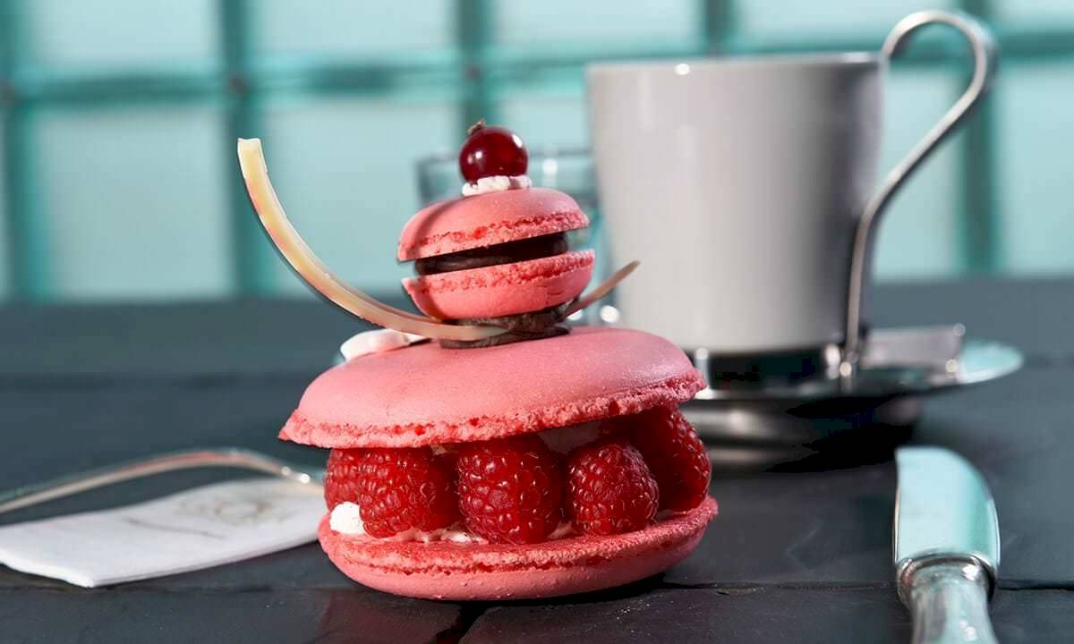 Ein rosa Marmorkuchen mit Himbeeren und einer Kirsche auf einem Tisch neben einer Tasse und einem Messer.