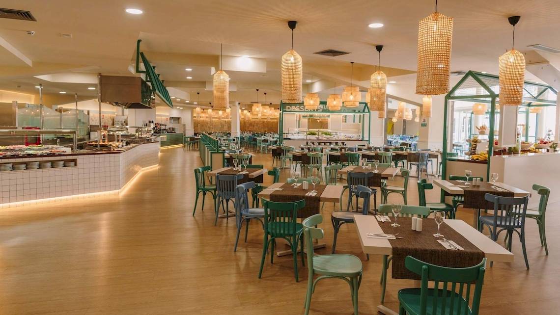 Innenansicht des Restaurants im Hotel Suites & Villas By Dunas mit vielen Tischen und Stühlen, Holzboden und Hängelampen.