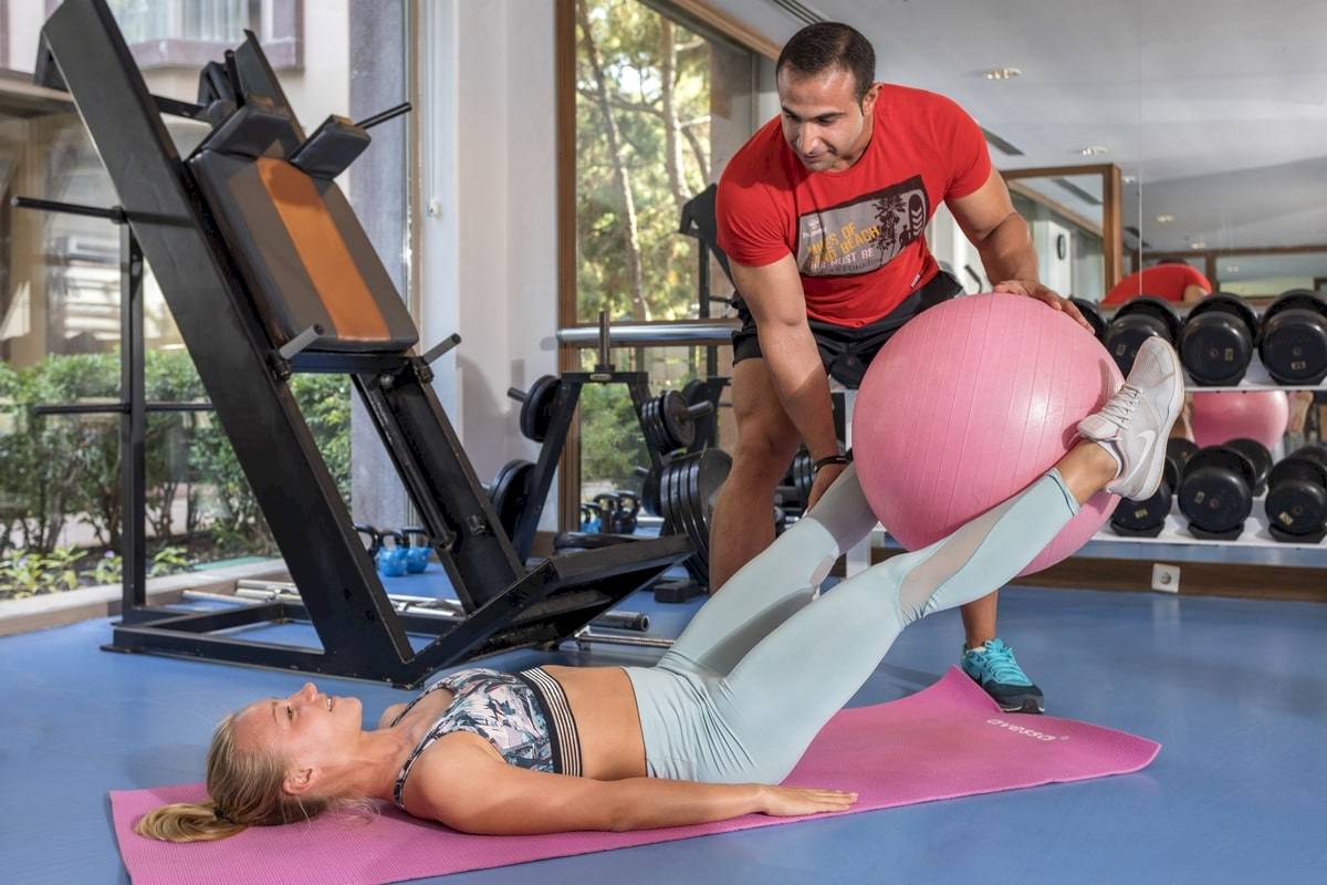 Fitnesstrainer im Fitnessstudio hilft einer Frau mit einem großen rosa Ball.