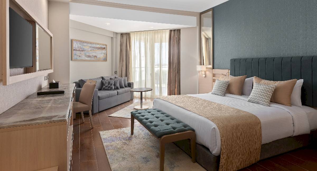 Innenansicht eines Hotelzimmers im Xanadu Makadi Bay mit einem großen Bett, einem Sofa, einem Fernseher und einem Schreibtisch.
