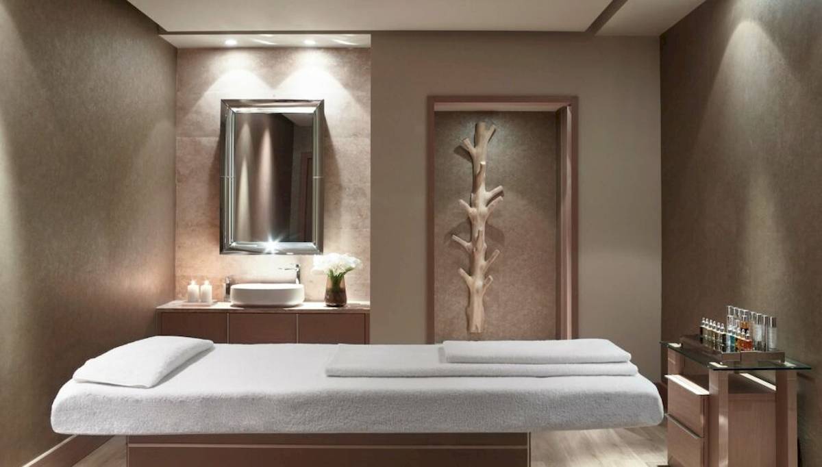 Ein eleganter Kosmetikbehandlungsraum im Ikos Dassia Hotel mit einem Behandlungsbett, einer Kommode und einem Spiegel.