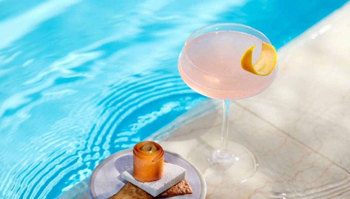 Ein Rosa Cocktail in einem Glas mit einer Zitronenscheibe steht am Rand eines Pools neben einem Teller mit einem gewickelten Keks und einem Marshmallow.