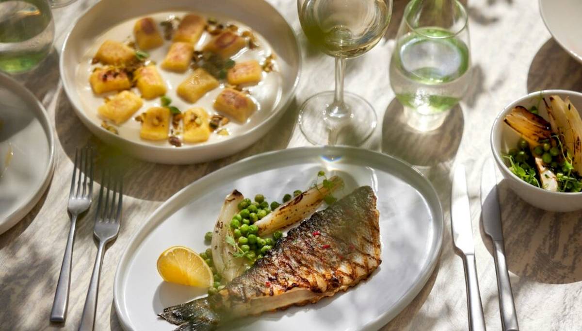 Ein Teller mit gegrilltem Fisch, Erbsen und Sellerie auf einem Tisch mit weiteren Gerichten und Getränken.