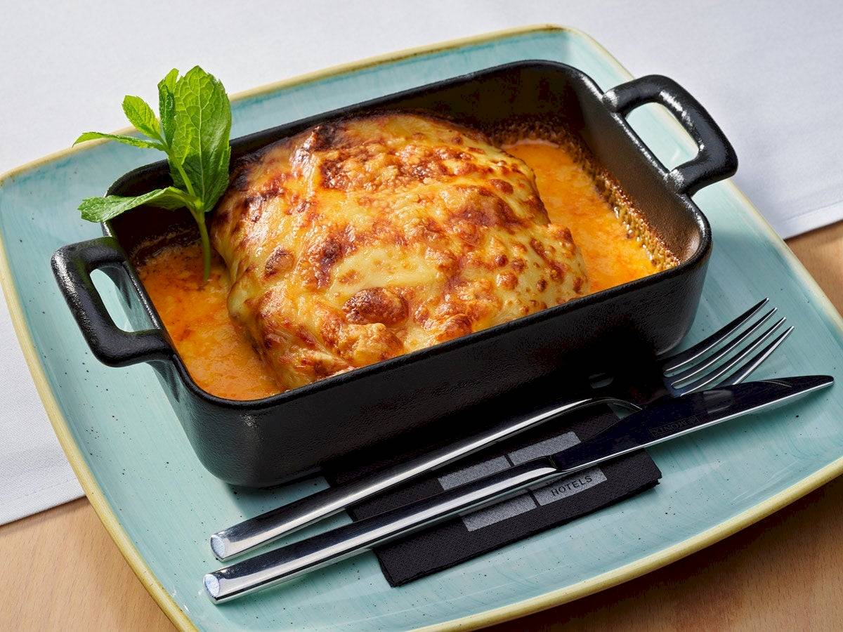 Lasagne im schwarzen Schälchen auf einem blauen Teller serviert, mit Messer, Gabel und Minzeblatt.