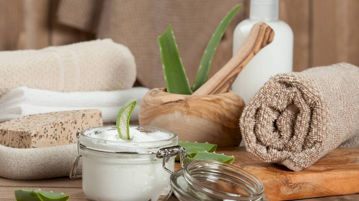 Ein Glas mit Aloe-Vera-Creme, umgeben von einem Holzmörser, einem Schwamm, einer Aloe-Vera-Pflanze und einem gerollten Handtuch.