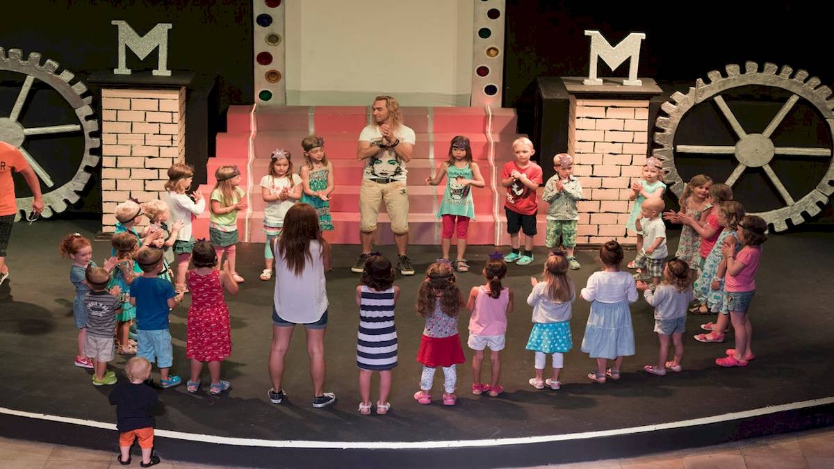 Gruppe von Kindern und einem Erwachsenen auf einer Bühne mit einem großen M im Hintergrund