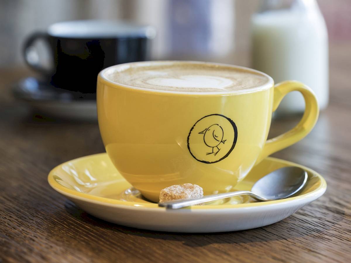 Ein gelber Kaffeebecher mit einem Vogeldesign auf einem blauen Unterteller und einer Tasse mit Milch.