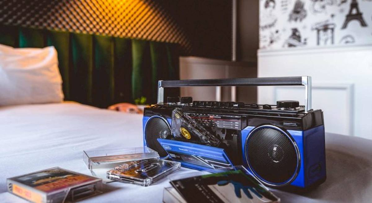 Ein Boombox auf einem Bett im ReMIX Hotel neben Musikkassetten und einem Kissen.