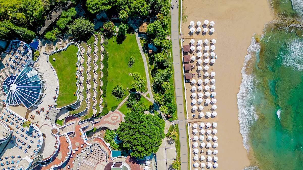 Luftaufnahme des Sonnenaufgang Queen Luxus Hotels mit Strand, weißen Sonnenschirmen und grüner Landschaft