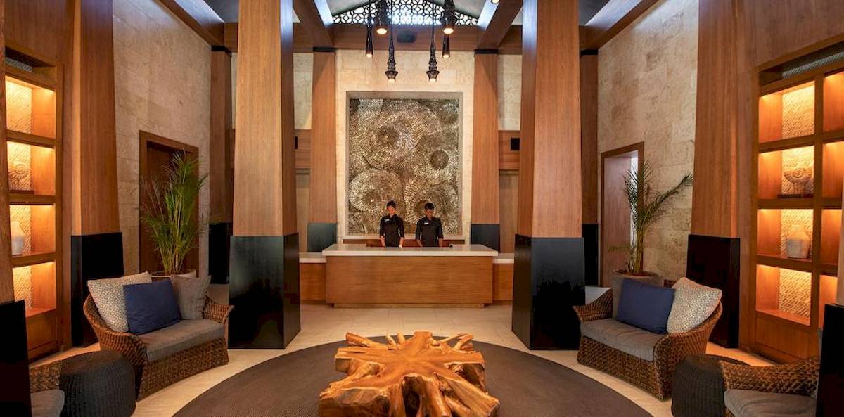 Hotellobby mit Holzelementen, modernen Möbeln, einem großen Holztisch und zwei Mitarbeitern hinter einer Rezeption.