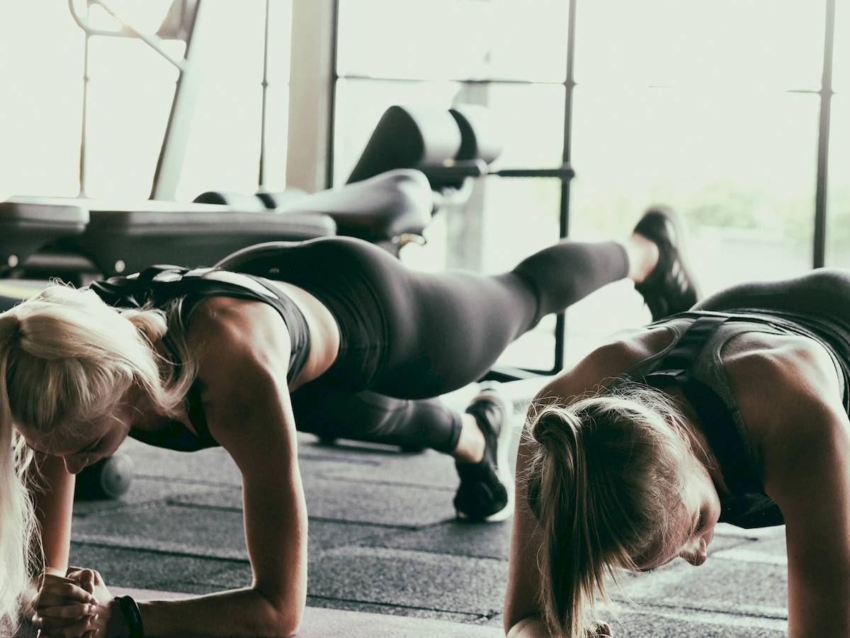 Zwei Frauen machen Push-Ups im Fitnessstudio mit Trainingsgeräten im Hintergrund.