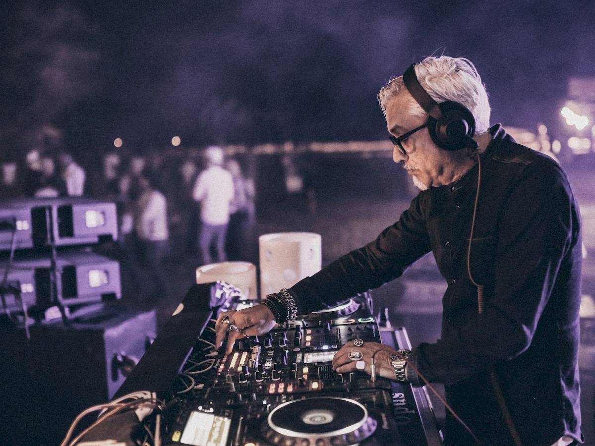 Ein älterer DJ mit weißer Frisur und schwarzer Kleidung spielt Musik in einer Nachtclub-Umgebung.