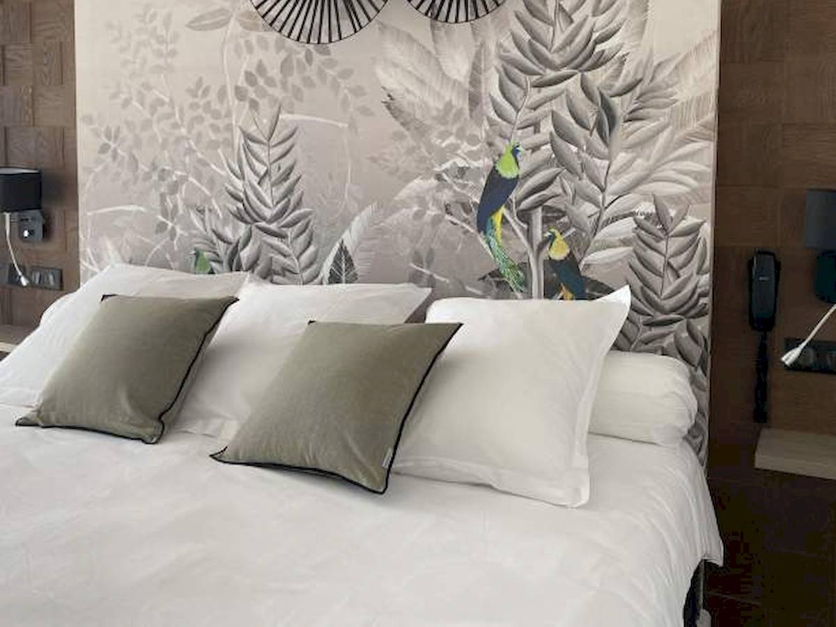 Ein Bett mit weißen Laken und grauen Kissen vor einer Wand mit exotischen Vogelmalereien im La Lagune Hotel.