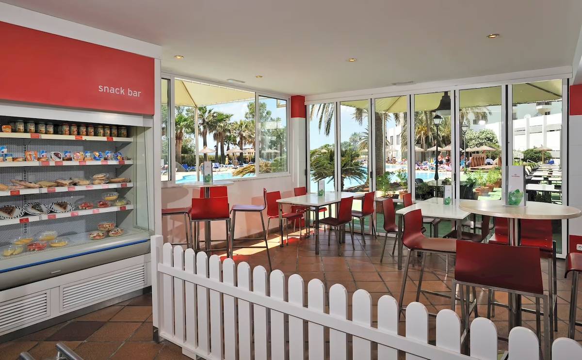 Innenansicht der Snackbar im Sol Milanos Pinguinos Hotel mit roten Stühlen und Tischen vor großen Fenstern.