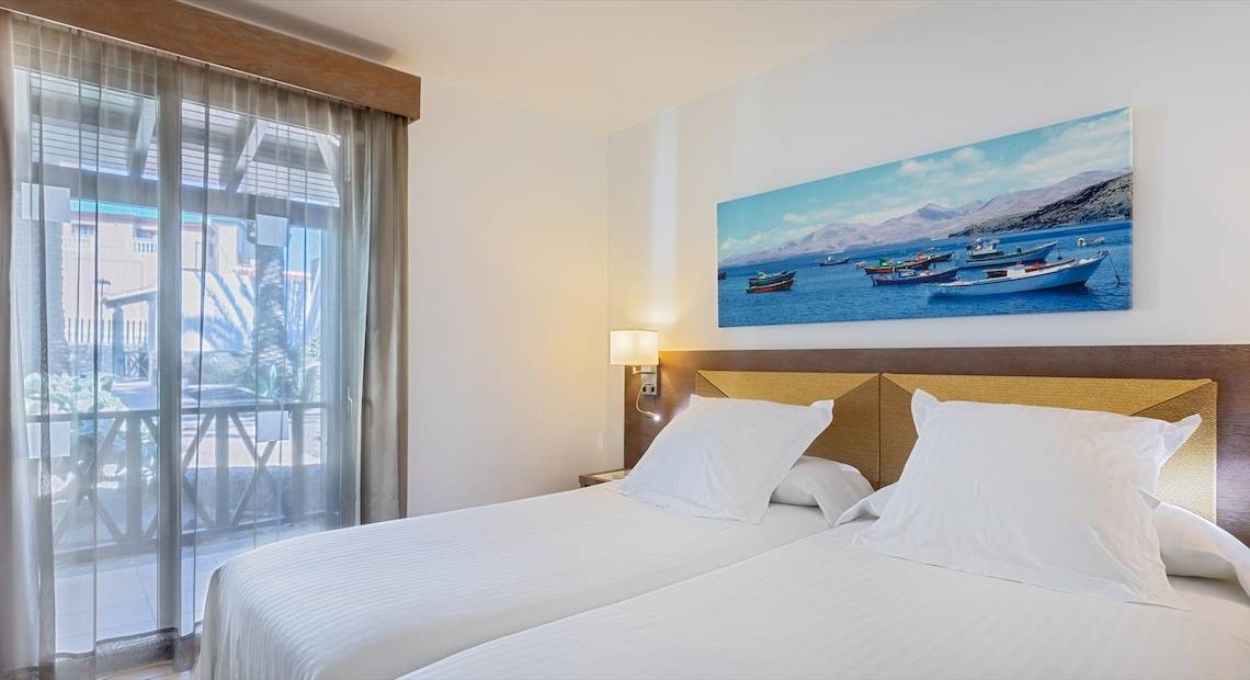 Doppelzimmer mit Meerblick im Barcelo Fuerteventura Castillo Hotel mit zwei Betten, weißem Dekor und einem Gemälde von Booten auf See.