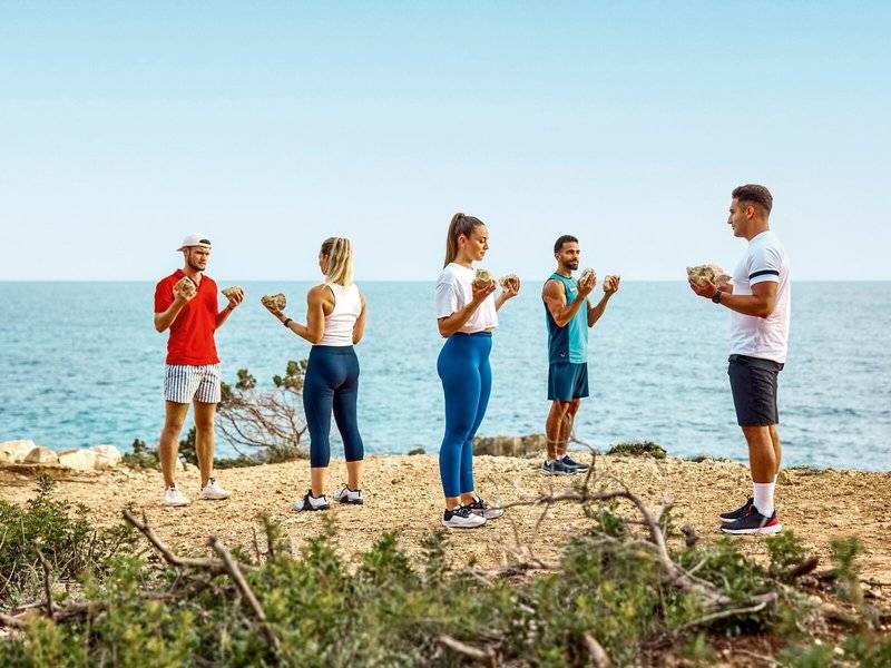 Gruppe von Menschen, die auf einem Strand Fitnessübungen mit Steinen machen, Victoria Menorca Hotel.