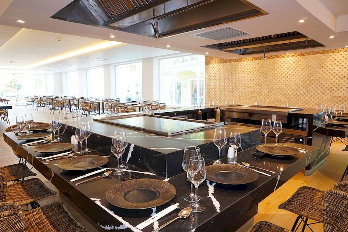 Innenansicht eines Restaurants mit einem langen Grilltisch, auf dem Teller, Gläser und Besteck bereitstehen.
