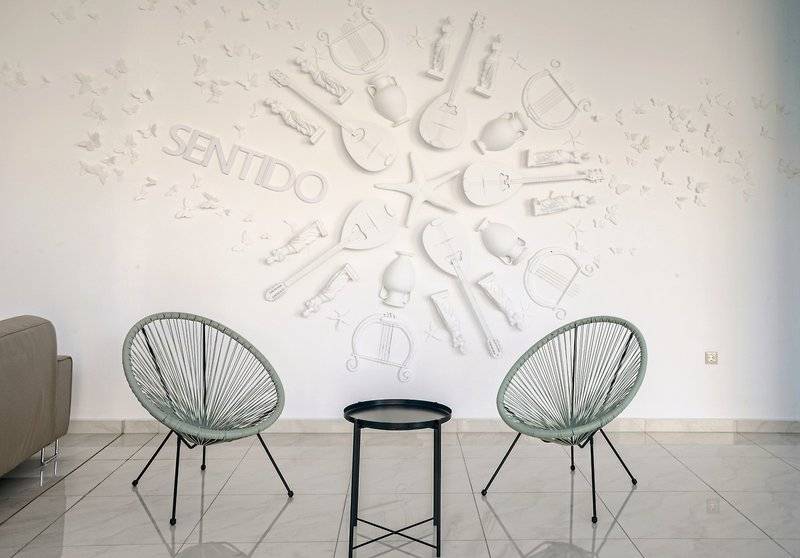 Eingangsbereich des Asterias Beach Resort mit einer Wand, die mit verschiedenen weißen Instrumenten und dem Wort 'Sentido' verziert ist.