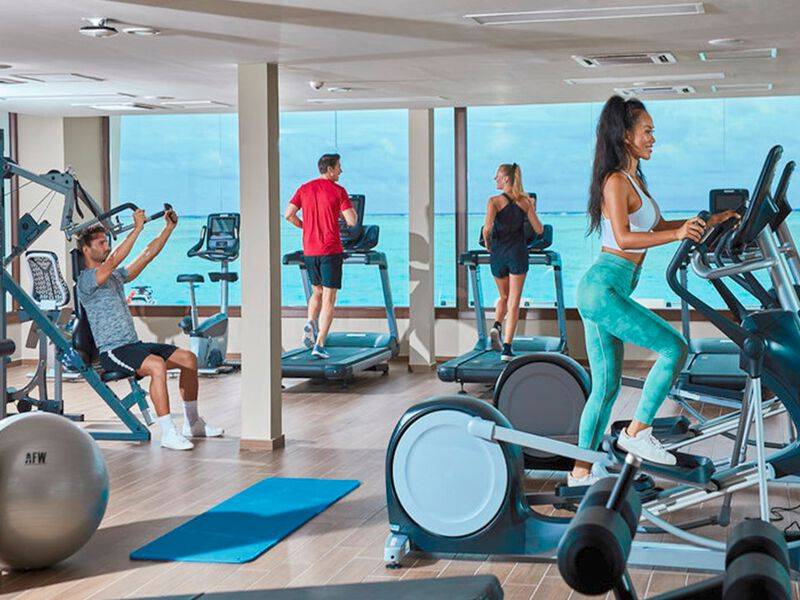 Fitnesscenter im RIU Atoll Hotel mit Mitgliedern, die auf Geräten trainieren und einen Blick auf das Meer genießen.
