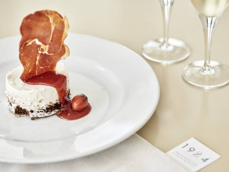 Ein Gericht auf einem weißen Teller serviert, dekoriert mit einem Stück Prosciutto, einer roten Soße und einer kleinen Tomate.