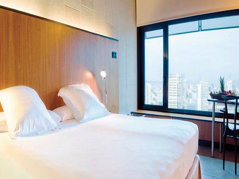 Ein modernes Hotelzimmer mit einem Bett, weißen Kissen, einer Lampe und einem Fenster mit Stadtblick im Hintergrund.