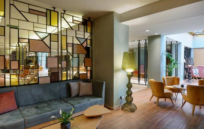 Sitzecke im Hotellobby von Dolce Athens Attica Riviera mit einem grauen Sofa, Kissen, und einer gelben Stehlampe.
