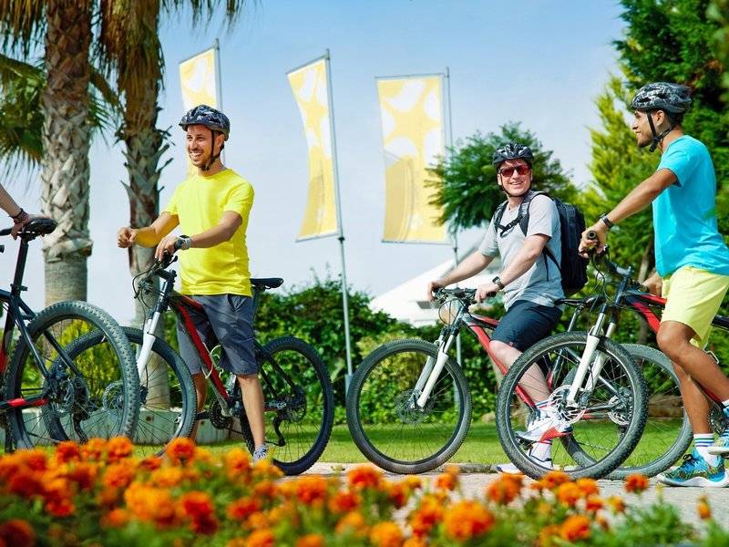 Drei Radfahrer im Magic Life Jacaranda, zwei in Helmen, einer mit Rucksack, auf gut gepflegtem Rasen.