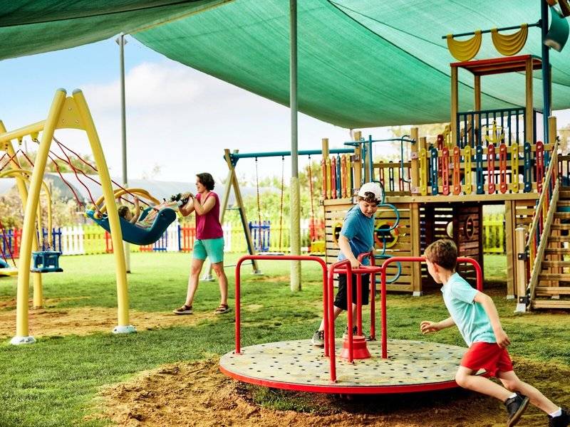 Familie auf dem Spielplatz im Magic Life Jacaranda Hotel, Kinder spielen auf Schaukeln und Karussell.