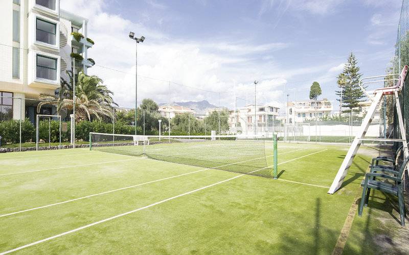 Tennisplatz vor dem UNAHOTELS Naxos Beach Sicilia Hotel mit Netz, Bänken und Gebäude im Hintergrund.