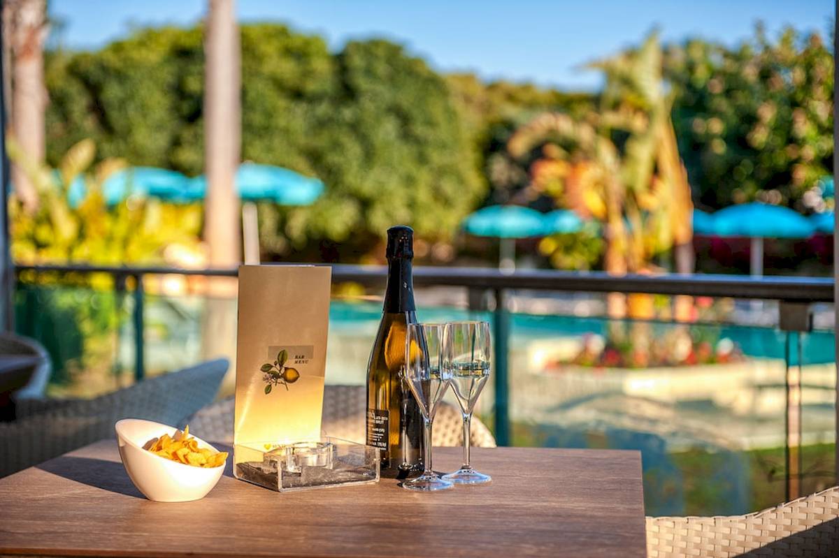 Terrasse im Acacia Palace Hotel mit einer Flasche und zwei Weinglaesern und einem Schwimmbad im Hintergrund
