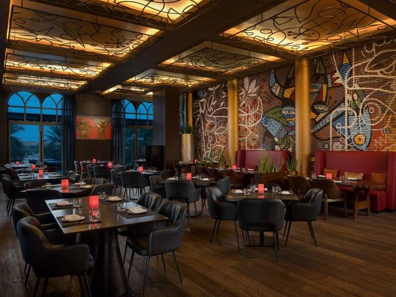 Innenbereich des Marsa Malaz Kempinski, The Pearl Restaurants mit geometrischen Wandgemälden und gedeckten Tischen.