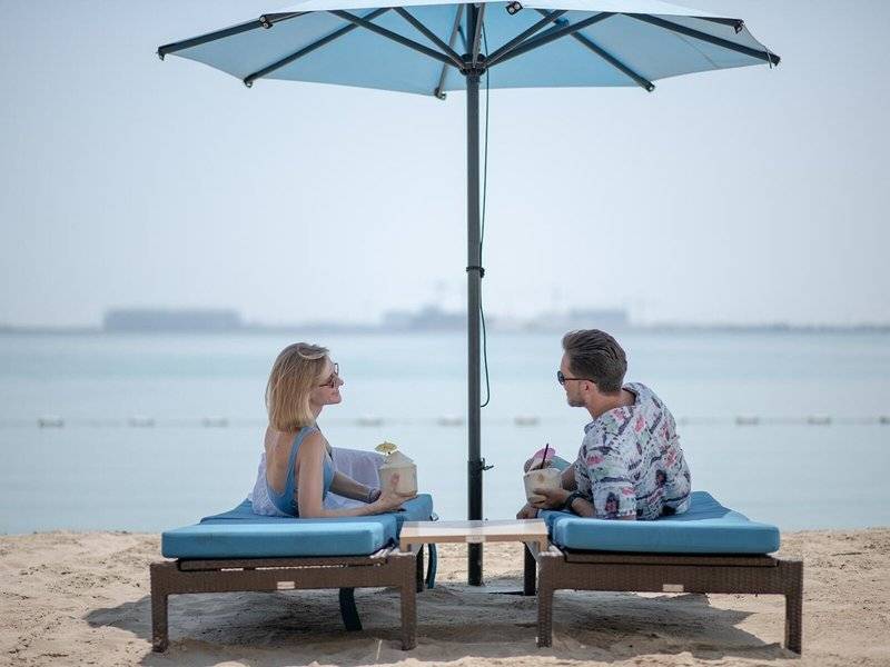 Ein Paar sitzt unter einem Schirm am Strand im Four Seasons Hotel Doha und genießt einen Drink.