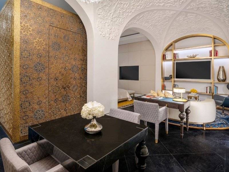Inneneinrichtung der Raffles Doha Suite mit goldenen Wandverkleidungen, einem schwarzen Tisch mit einer Blumenvase, einem Schreibtisch, Sesseln und einem Fernseher.