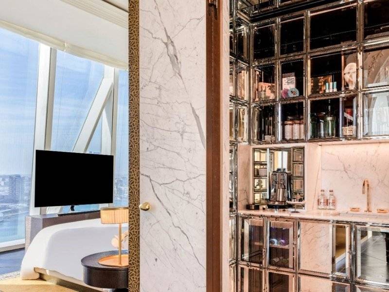 Innenansicht des Schlafzimmers im Raffles Doha Hotel mit Marmorwänden, einem großen Bett, Fernseher und Minibar.
