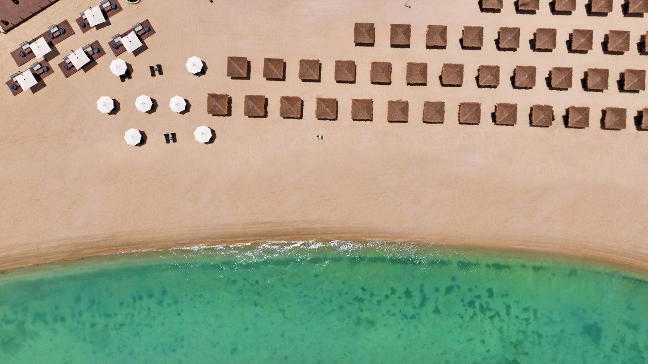 Luftaufnahme eines Strandes mit vielen braunen Strohhütten und weißen Sonnenschirmen.