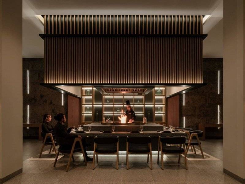 Das Interieur des JAZ Riviera Restaurants mit einem Koch am offenen Feuer, umgeben von Gästen an einem langen Tisch.