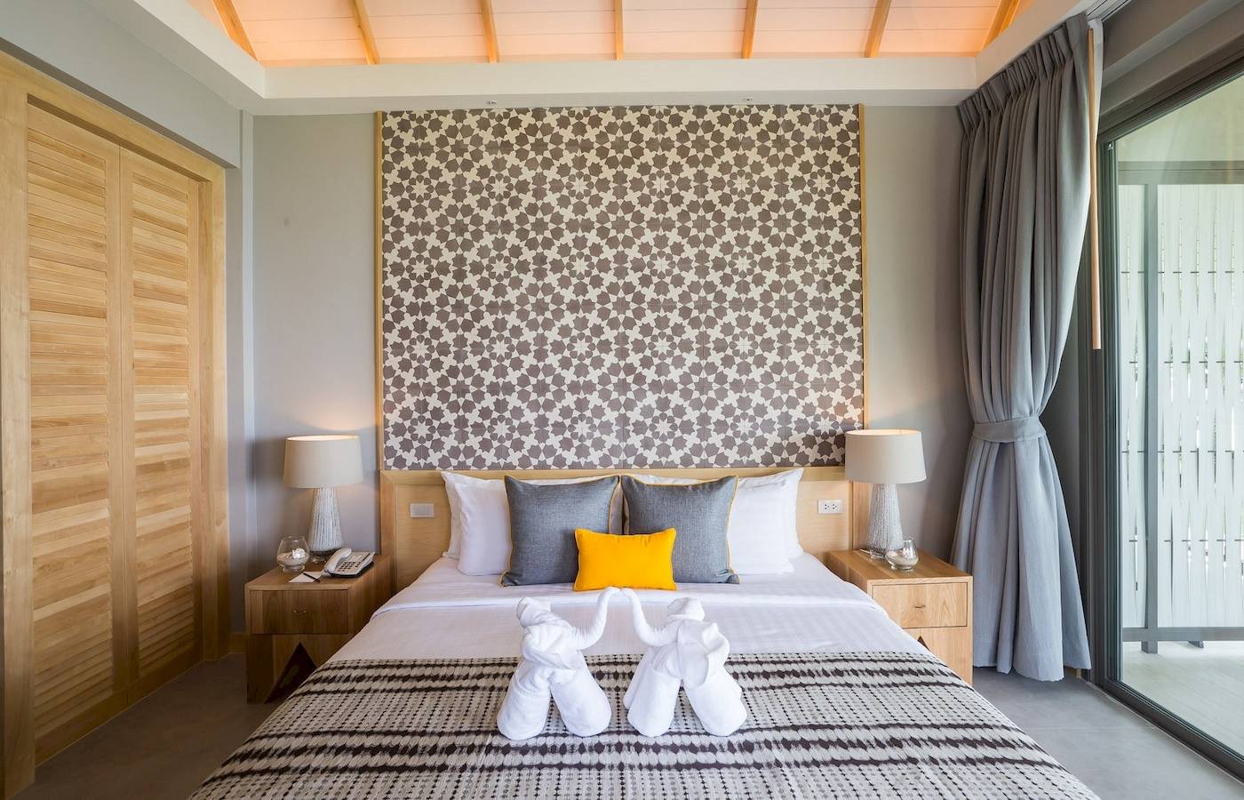 Schlafzimmer im KC Grande Resort Koh Chang mit gemusterter Wand, zwei Nachttischen, Lampen, Telefon und zwei weißen Elefanten-Handtüchern.