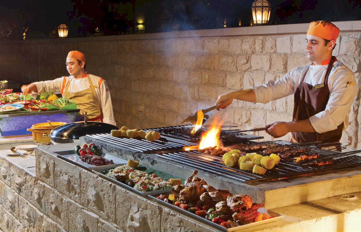 Grill im Traders Hotel Qaryat Al Beri mit Köchen, die Fleisch und Gemüse zubereiten.