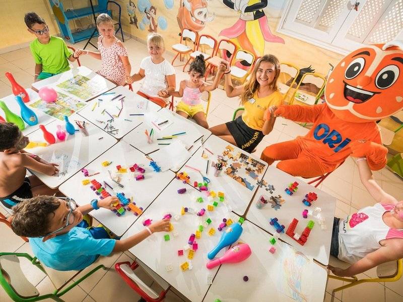 Kinder im Hotel 'Iliade Djerba by Magic Hotels' nehmen an einer Bastelaktivität teil, mit bunten Bausteinen und Stiften auf Tischen.