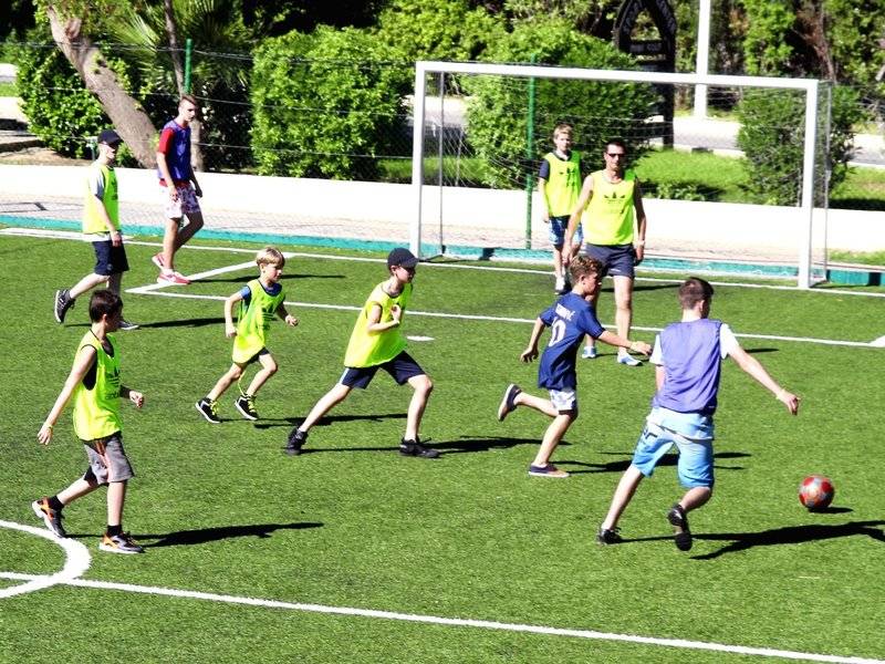 Kinder in neonfarbenen Trikots spielen Fußball auf einem Feld mit einem Tor im Hintergrund.
