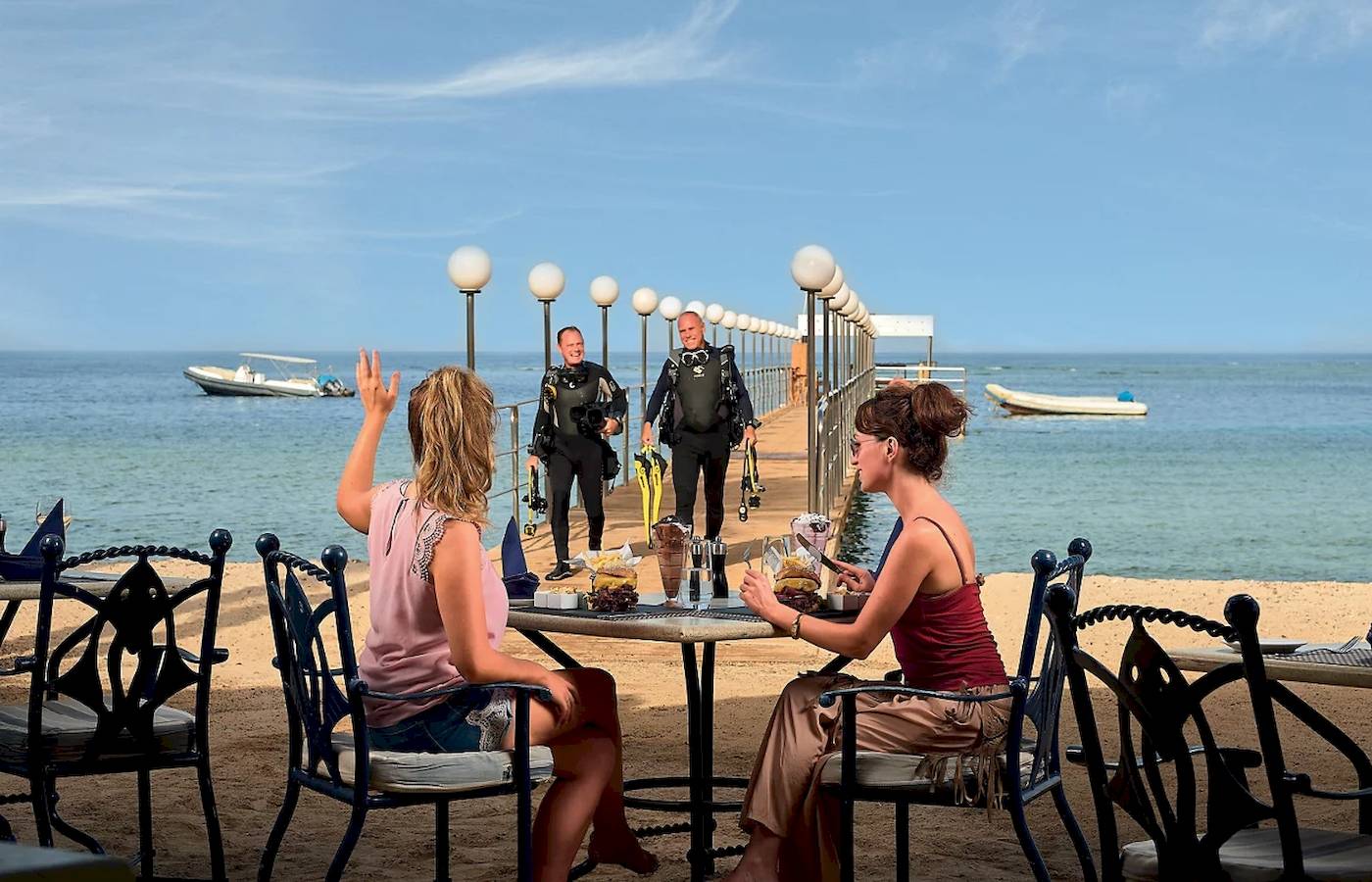 Scuba-Taucher, die auf einem Steg des Mövenpick Resort El Quseir stehen, während zwei Frauen am Tisch sitzen und essen.