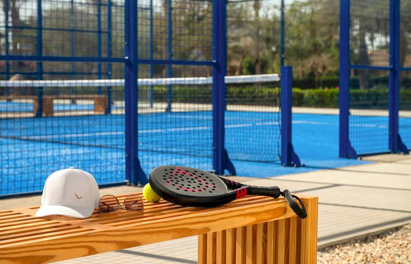 Tennisschläger, Ball, Hut und Sonnenbrille auf einer Holzbank neben einem Tennisplatz