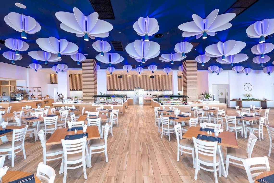 Innenansicht des Restaurants im Arina Beach Resort mit weißen Tischen, Stühlen und blauer Decke mit Beleuchtung.