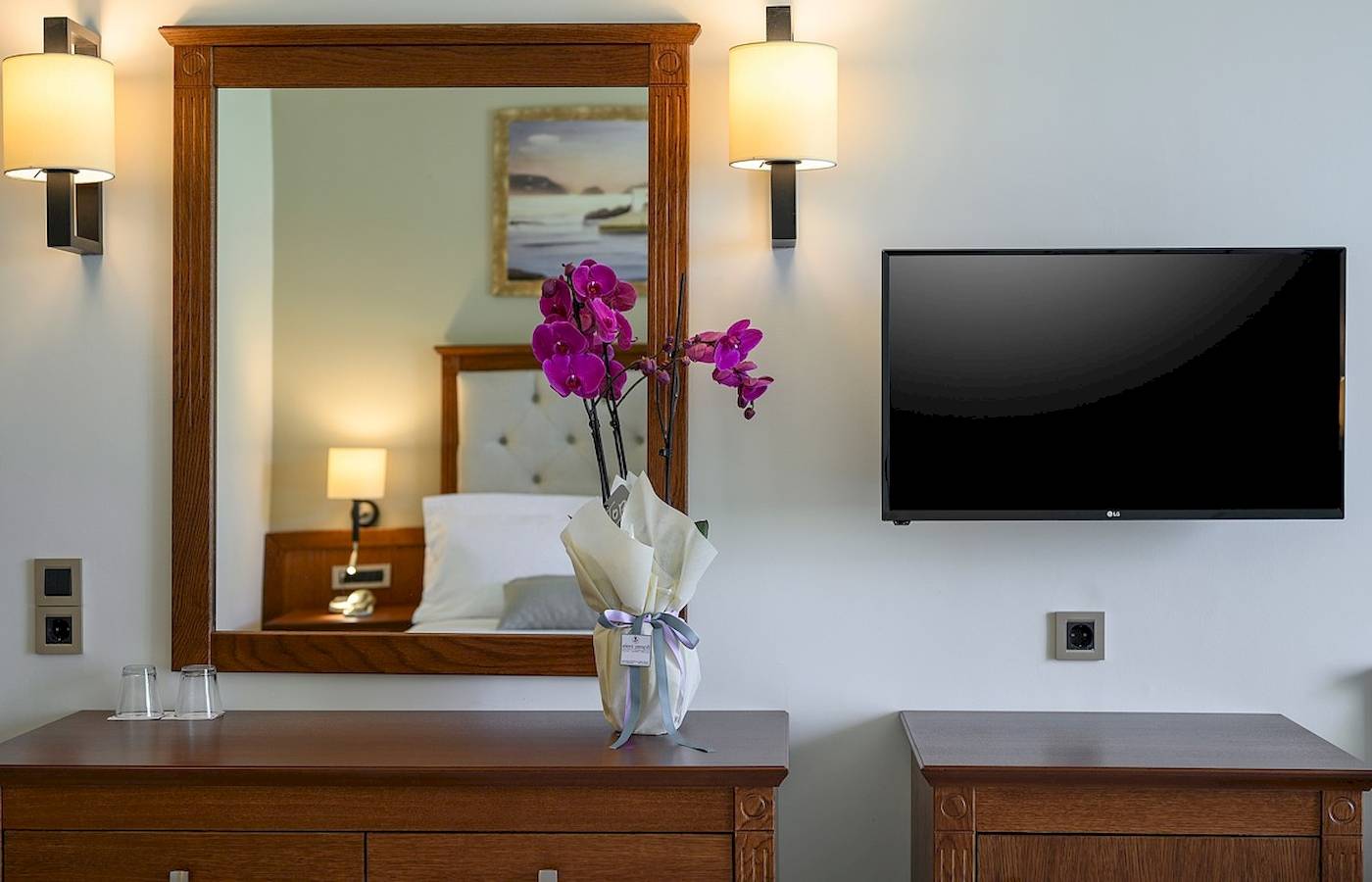 Ein Hotelzimmer mit einem Spiegel und einem Fernseher an der Wand, neben einem Nachttisch mit Blumen.