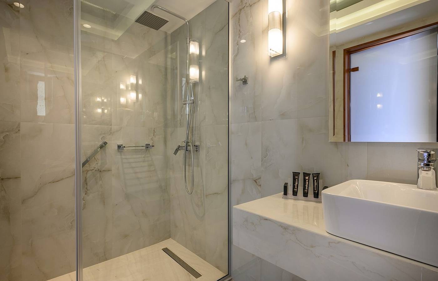 Ein elegantes Badezimmer im Vantaris Palace Hotel mit Marmorwänden, einer Glasdusche und einem Waschbecken.