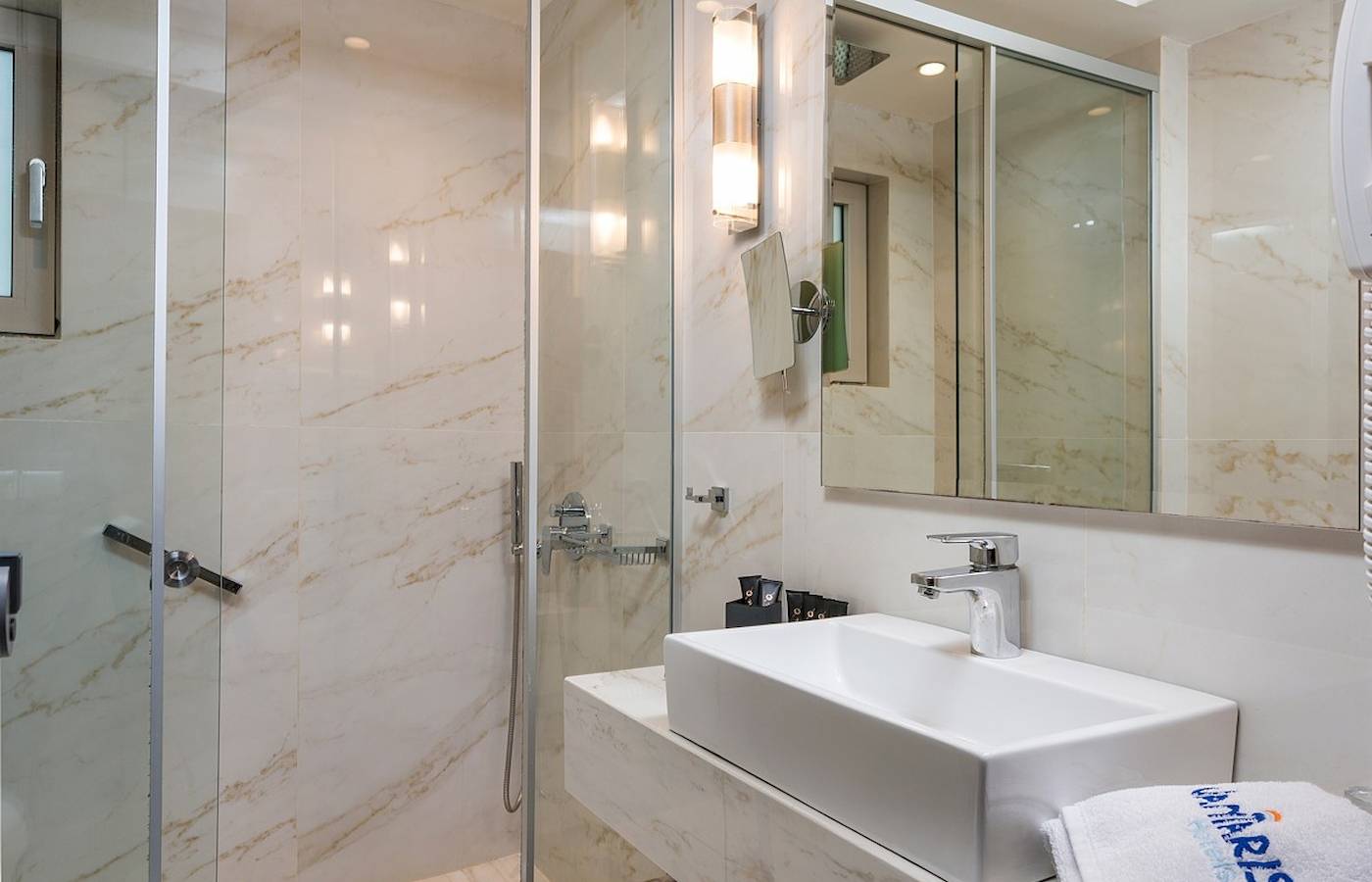Badezimmer im Vantaris Palace Hotel mit Marmorwänden, Glasdusche und Waschbecken.